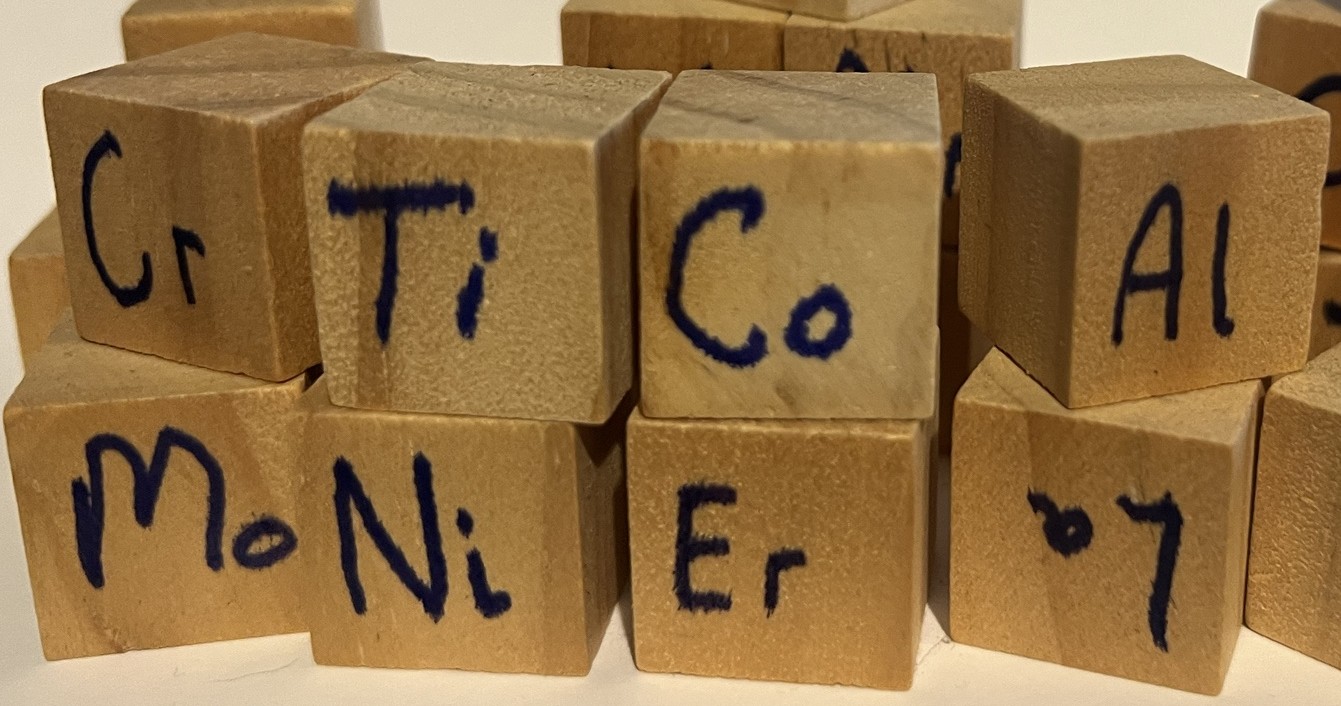 Wooden building blocks hand-labelled with chemical symbols, spelling out CRITICAL MINERAL: Cr Ti Co Al over Mo Ni Er and an upturned La